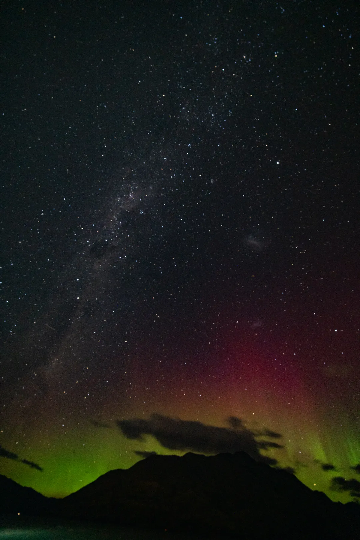 A vertical night sky photograph showing the bright band of the Milky Way galaxy stretching down towards a silhouetted mountain range. Low on the horizon, the vibrant green and deep magenta curtains of the Aurora Australis glow brightly behind dark clouds. The entire sky is filled with thousands of sharp, clear stars.