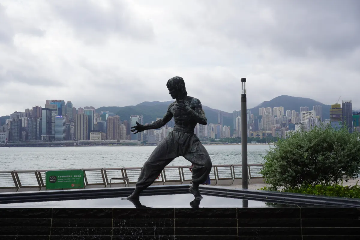 A bronze statue of martial artist Bruce Lee in a classic shirtless combat stance, positioned over a shallow, dark-tiled water fountain. In the background, the dense skyline of Hong Kong Island stretches across Victoria Harbour under an overcast, cloudy sky, with green mountains rising behind the high-rises.