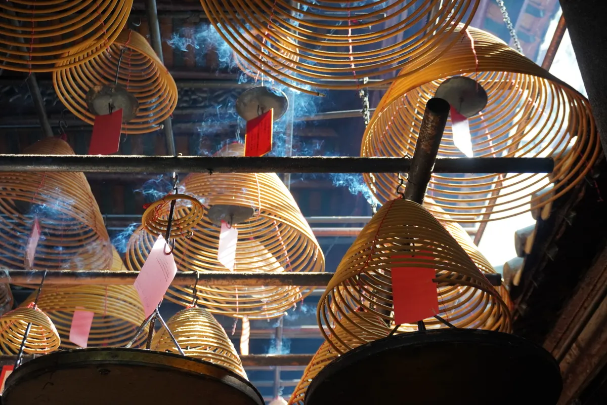 A low-angle close-up shot looking up at several large, cone-shaped spiral incense coils hanging from the ceiling of a dimly lit temple. Light streams from above, illuminating the rising trails of blue-grey smoke. Small, bright red and pink rectangular paper tags are suspended from the center of each yellow-orange coil, set against a dark background of wooden roof beams.