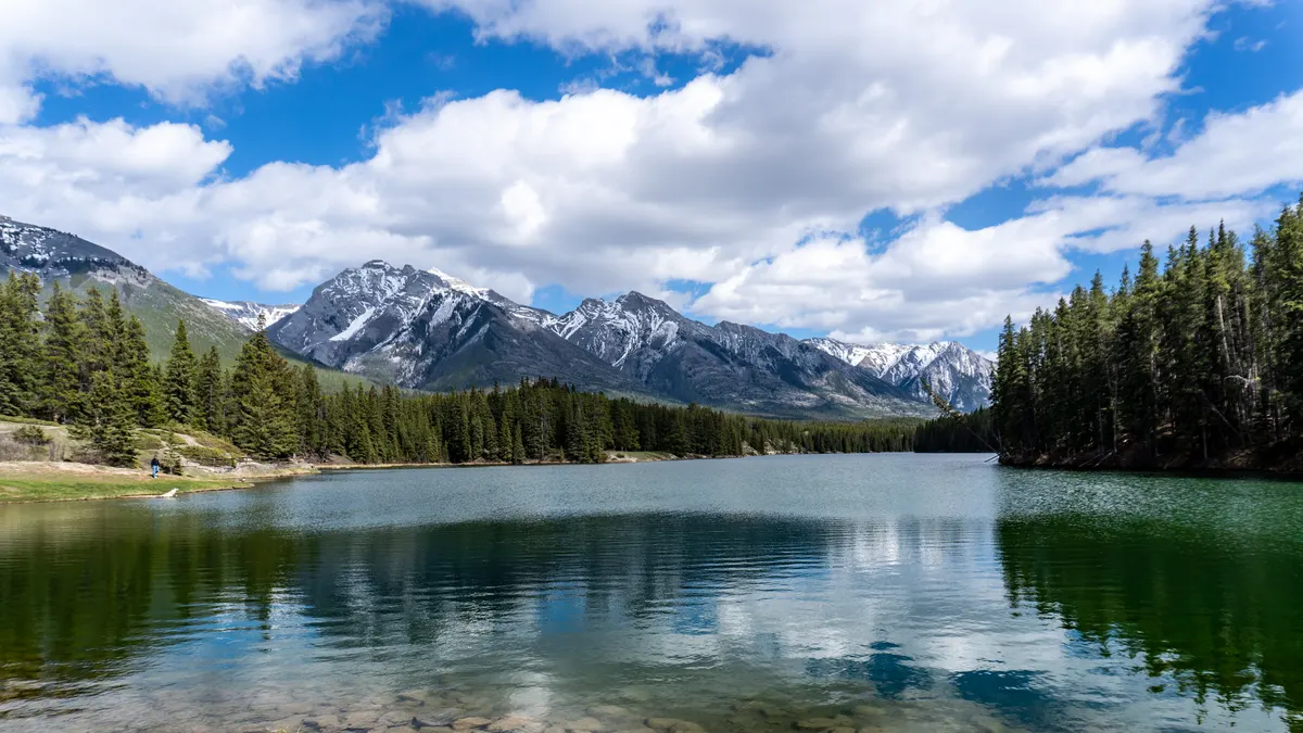 A wide landscape photograph of a calm, clear alpine lake reflecting a deep green forest along its banks. In the far background, a large range of rugged, snow-capped mountains stands underneath a bright blue sky filled with puffy white clouds. The shallow water in the immediate foreground reveals smooth rocks beneath the surface.
