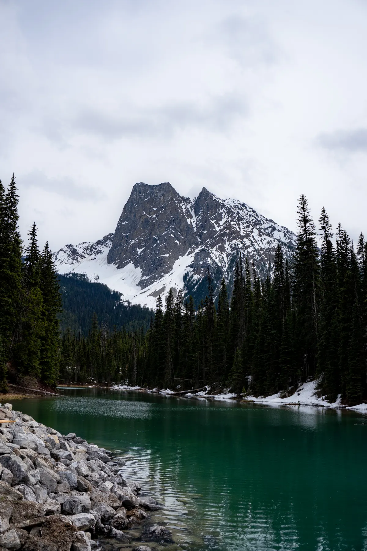 A vertical landscape photograph of a vibrant, emerald-green lake surrounded by a dense forest of tall, green pine trees. In the bottom left foreground, a grey rock wall lines the edge of the water. In the background, the massive, jagged peak of Mount Burgess rises with patches of white snow under a cloudy, overcast sky.