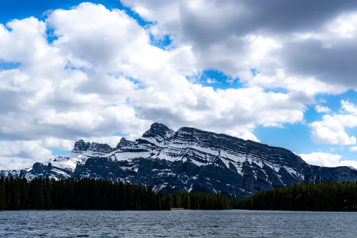 A wide horizontal landscape shot looking across the rippling waters of Two Jack Lake. A thick, dark green pine forest lines the far shore, above which the wide, dramatic peaks of Mount Rundle rise with patches of white snow. The sky above is filled with dramatic, puffy white clouds and patches of bright blue.