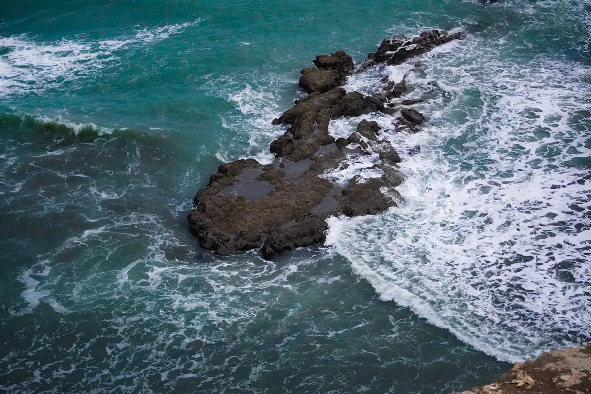 A high-angle, top-down shot of a long, dark, jagged volcanic rock shelf extending into a turquoise ocean. Waves crash violently against the rock formation, creating intricate patterns of white foam and sea spray that swirl around the stone. The deep teal water shows shifting textures from the turbulent swells.