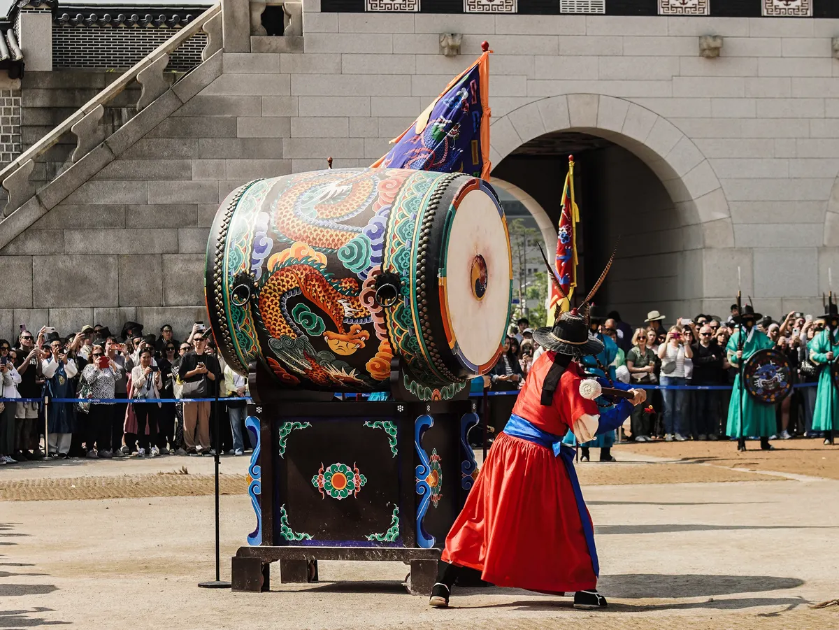 A traditional Korean royal guard in a bright red robe and black brimmed hat strikes a large, ornately decorated ceremonial drum. The drum features vibrant painted details of a golden dragon amidst swirling blue and green clouds. In the background, a crowd of tourists watches from behind a blue barrier in front of the stone arched gateway of Gyeongbokgung Palace under bright daylight.