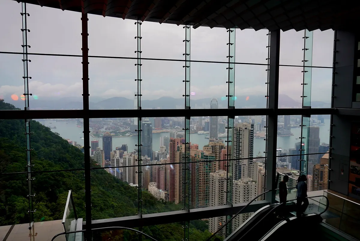 A wide interior view looking out through a massive, multi-paneled glass window wall from inside a modern building on a hillside. Two silhouetted figures stand near an escalator in the lower right foreground. Outside, the dense skyscraper skyline of Hong Kong spreads out across the waterfront of Victoria Harbour under a hazy, overcast sky, flanked by a steep, lush green forested hill on the left.